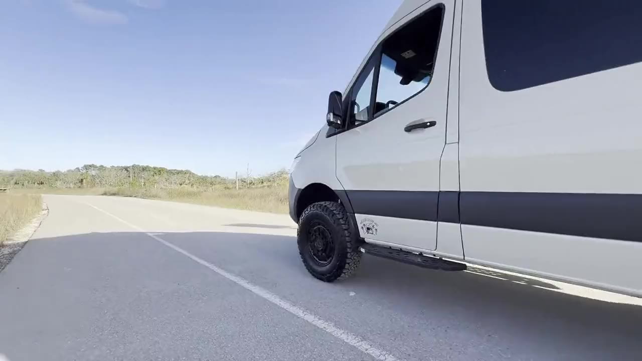 A white Sprinter camper van driving away down a coastal road