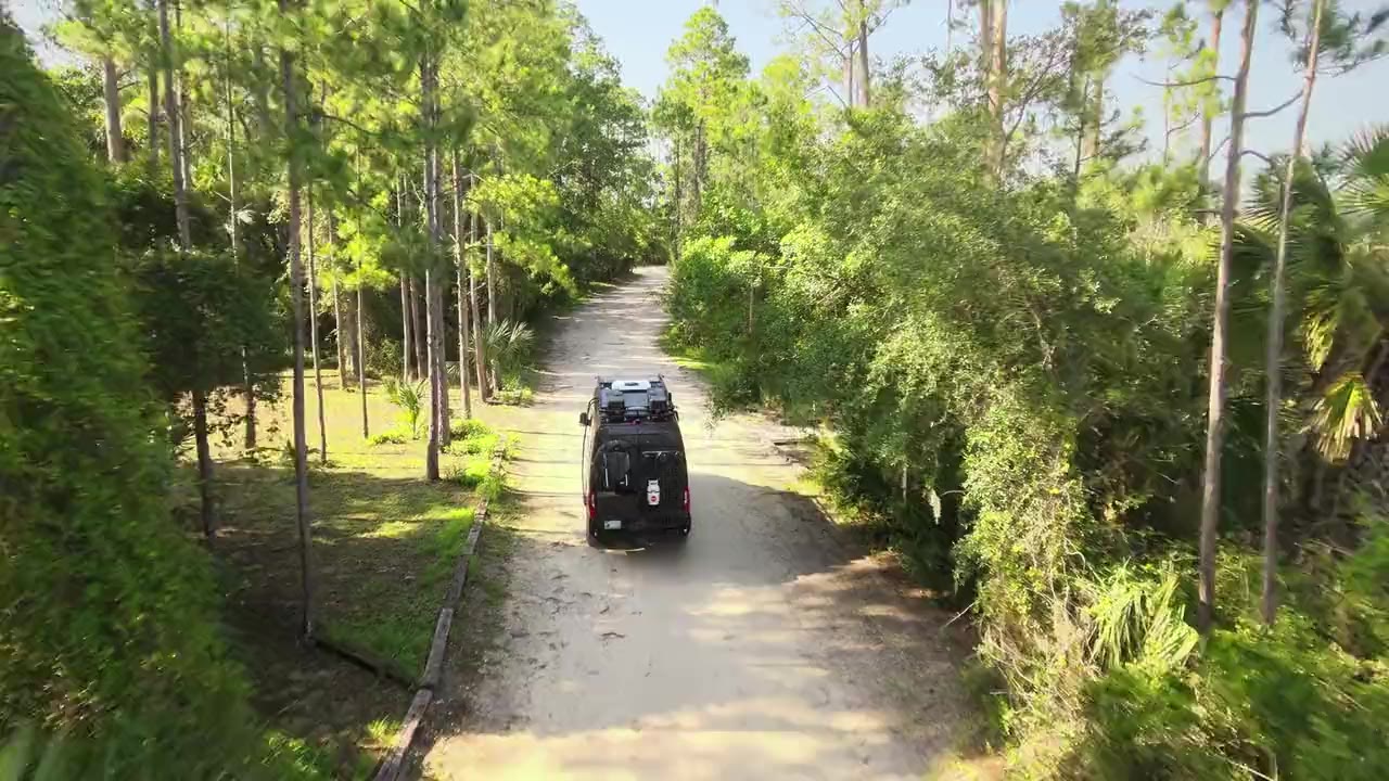 A dark Sprinter camper van driving down a sandy forest road