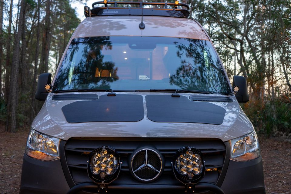 A sand-colored Sprinter with off-road lights and a roof rack