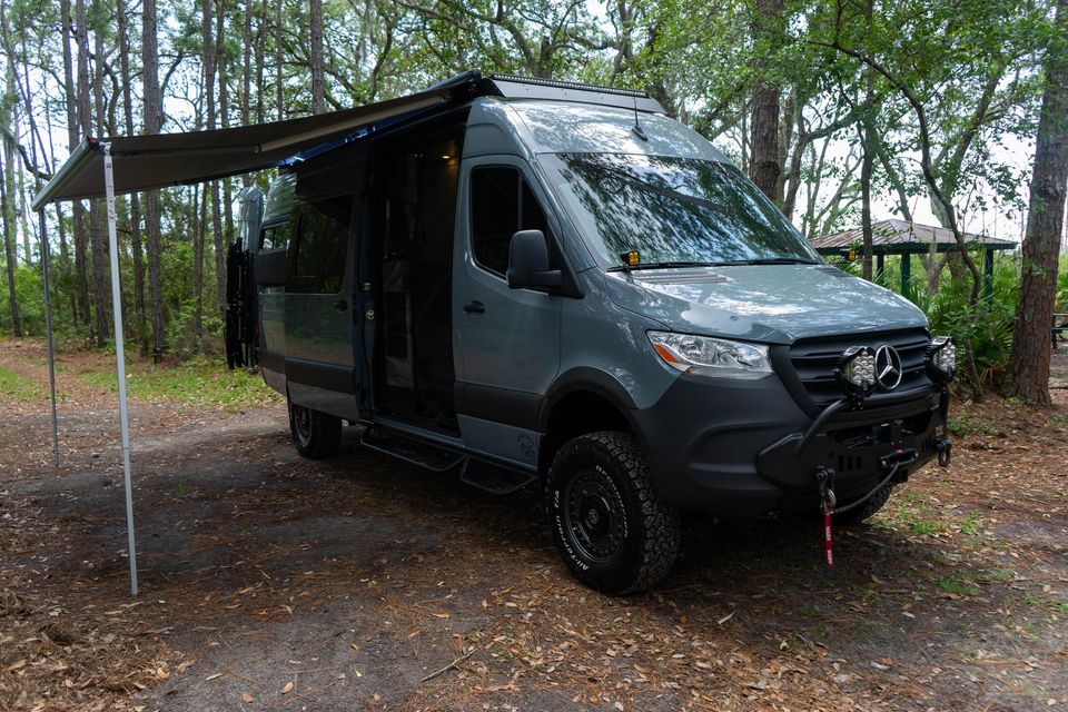 A dark Sprinter with its awning out in the woods