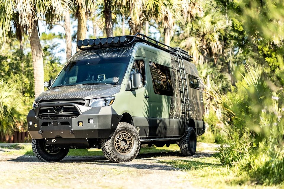 A green Sprinter with an off-road bumper and lights among palms